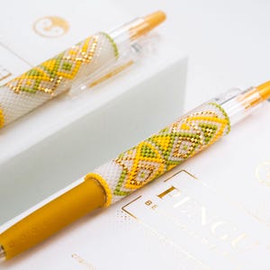 Op de afbeelding: Twee pennen met gele, groene en gouden kralenpatronen op een witte achtergrond. De pennen hebben gele inktpunten en liggen op een witte doos met de tekst "PENGU BEADING JEWELRY". Een uniek en kleurrijk schrijfactie.