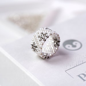 May include: A white and silver beaded ring with a geometric pattern. The ring is on a white surface with a yin yang symbol and the word "Peace" printed on it.