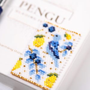 Könnte beinhalten: Ein Perlen-Lesezeichen mit einem Blumen- und Ananas-Design in Blau, Gelb und Gold. Das Lesezeichen befindet sich auf einem weißen Hintergrund, auf dem das Wort "PENGU" zu sehen ist. Das Lesezeichen ist eine charmante Sache.