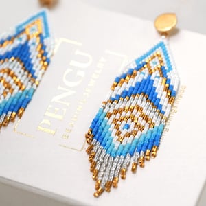 May include: A pair of blue, white and gold beaded dangle earrings. The earrings are made with small, colourful beads and have a geometric pattern. The earrings are hanging from gold hooks.