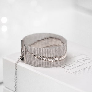 K&ouml;nnte beinhalten: Ein silbernes Perlenarmband mit einer Kette und einem silbernen Verschluss. Das Armband hat ein geometrisches Muster mit einem wei&szlig;en Streifen. Das Armband liegt auf einer wei&szlig;en Schachtel mit dem Text "PENGU Beading Jewels".