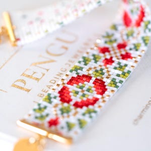 Könnte beinhalten: Ein Perlenarmband mit weißer Basis, das ein sich wiederholendes Muster aus roten und grünen geometrischen Formen aufweist. Das Armband hat goldfarbene Verschlüsse und Akzente. Der Hintergrund enthält den Text "PENGU BEADING JEWELRY".