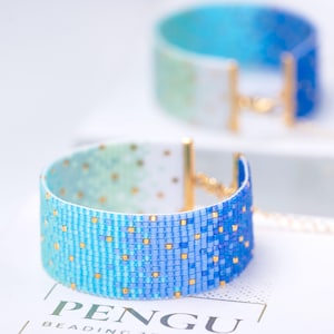 May include: Two beaded bracelets with a gradient of colors from white to blue. The bracelets have gold-colored clasps. The lower bracelet has the word "PENGU" visible. The bracelets are made of small, square beads.