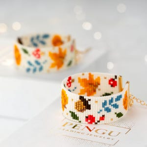 May include: Two white beaded bracelets with a colourful autumn leaf pattern. The bracelets are on a white surface with the text "PENGU BEADING JEWELRY".