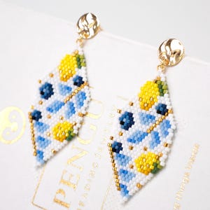 Puede incluir: Un par de pendientes de cuentas con un diseño geométrico. Los pendientes tienen forma de diamante con cuentas blancas, azules, amarillas y doradas. Los pendientes tienen postes dorados.