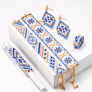 Könnte beinhalten: Eine Sammlung von Schmuck und Accessoires mit Perlenbesatz und einem geometrischen Muster in Blau, Weiß und Gold. Das Set umfasst Armbänder, Ohrringe, einen Ring und einen Stift. Der Schmuck wird auf einer weißen Box präsentiert.