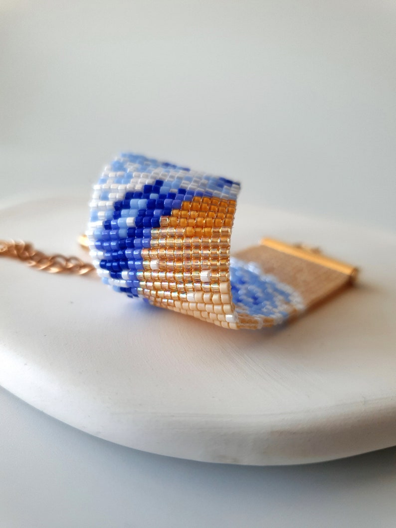K&ouml;nnte beinhalten: Ein Armband aus Perlen mit einem geometrischen Muster in Blau, Wei&szlig; und Gold. Das Armband ist aus kleinen, bunten Perlen gefertigt und hat einen goldenen Verschluss.