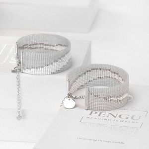 K&ouml;nnte beinhalten: Zwei silberne Perlenarmb&auml;nder mit einem wei&szlig;en und grauen geometrischen Muster. Die Armb&auml;nder haben einen silbernen Verschluss und einen kleinen silbernen Anh&auml;nger mit dem Wort "PENGU" und dem Text "BEADING JEWELRY" darauf. Die Armb&auml;nder liegen auf einer wei&szlig;en Oberfl&auml;che mit dem Text "Charming things inside" darauf.