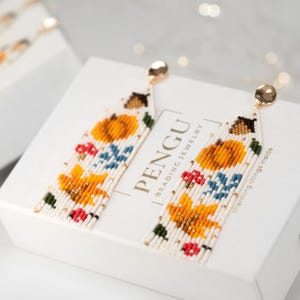 May include: A pair of long, beaded earrings with a fall-themed design. The earrings feature a white background with orange pumpkins, red berries, green leaves, and yellow flowers. The earrings are made with small, colourful beads and are attached to gold-coloured hooks.