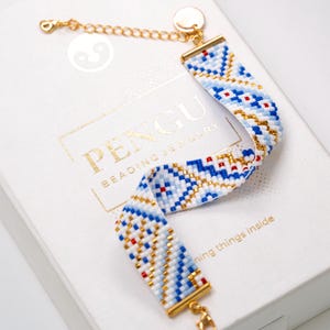 Peut inclure: Un bracelet en perles blanches et bleues avec des accents dorés et un fermoir doré. Le bracelet est sur une boîte blanche avec le texte "PENGU BEADING JEWELRY" et "ning things inside".