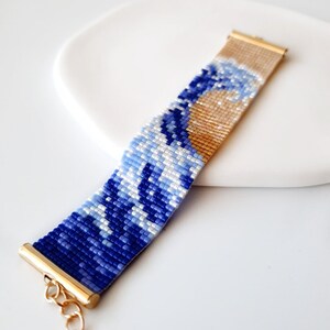 K&ouml;nnte beinhalten: Ein blaues und wei&szlig;es Perlenarmband mit einem goldenen Verschluss, das ein Wellenmuster darstellt.