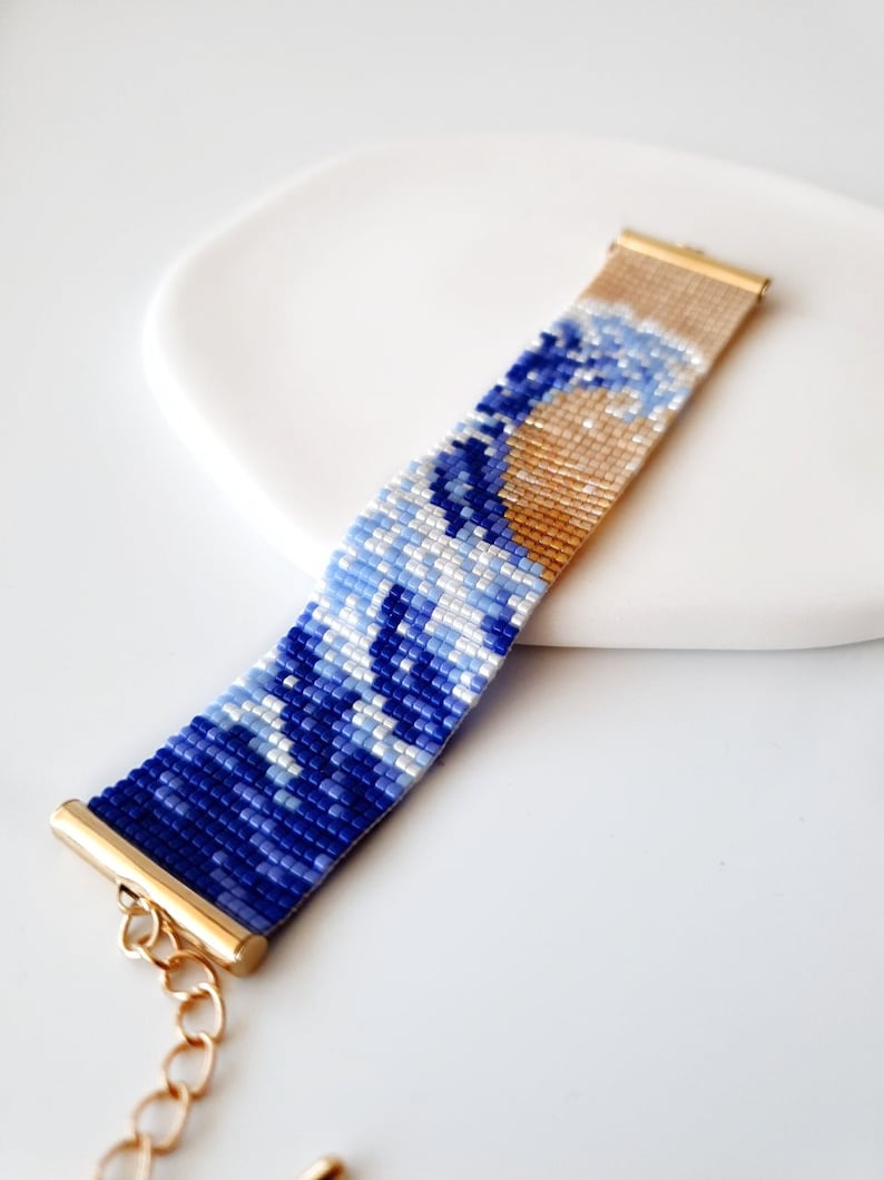 K&ouml;nnte beinhalten: Ein blaues und wei&szlig;es Perlenarmband mit einem goldenen Verschluss, das ein Wellenmuster darstellt.