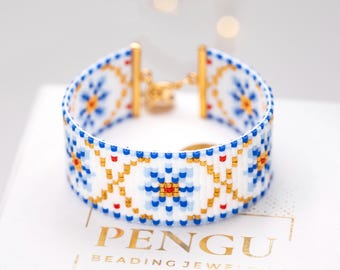 May include: A white beaded bracelet with a blue and gold floral pattern. The bracelet has a gold clasp and is displayed on a white surface. The text "PENGU BEADING JEWELRY" is visible.