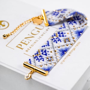 Könnte beinhalten: Ein weißes und blaues Perlenarmband mit goldenen Akzenten. Das Armband hat einen Verschluss und liegt auf einer weißen Schachtel mit dem Text "PENGU BEADING JEWELRY" und "charming things inside".