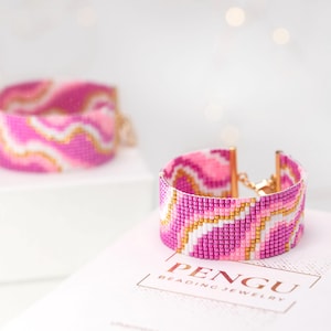 May include: Two pink and white beaded bracelets with a gold accent. The bracelets have a wavy pattern and are stacked on top of each other. The bracelets are on a white surface with the text "PENGU BEADING JEWELRY" printed on it.