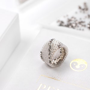 Peut inclure: Une bague en perles argentées et blanches avec un motif géométrique. La bague est posée sur une surface blanche avec un petit logo.