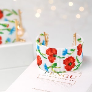Peut inclure: Un bracelet manchette blanc perlé avec un motif floral de coquelicots rouges, de fleurs bleues et de feuilles vertes. Le bracelet a des fermoirs dorés et est présenté sur une surface blanche. La marque "PENGU BEADING JEWELRY" est visible.