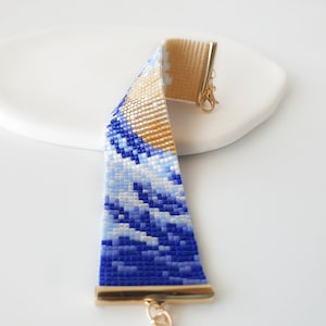 K&ouml;nnte beinhalten: Ein blaues und goldenes Perlenarmband mit Wellenmuster. Das Armband hat einen goldenen Verschluss und eine goldene Kette.