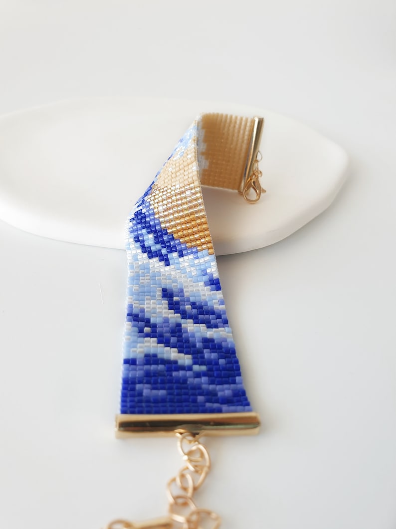 K&ouml;nnte beinhalten: Ein blaues und goldenes Perlenarmband mit Wellenmuster. Das Armband hat einen goldenen Verschluss und eine goldene Kette.