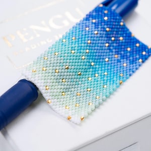 May include: A beaded bookmark with a gradient of white, teal, and blue, accented with small gold beads. The bookmark is rectangular and appears to be handmade. A blue pen is inserted into the bookmark. The word "PENGU" is visible on the background.
