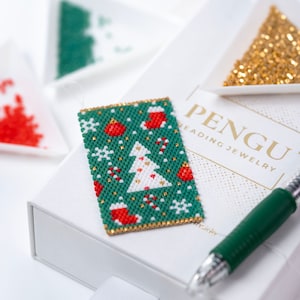May include: A close-up of a green, white, and red beaded Christmas tree ornament. The ornament is on a white box with the text "PENGU HEADING JEWELRY".
