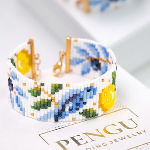 May include: A white beaded bracelet with a floral pattern in yellow, blue, green, and purple. The bracelet has gold-colored clasps and a chain. The bracelet is on a white surface with the brand name "PENGU" in gold.