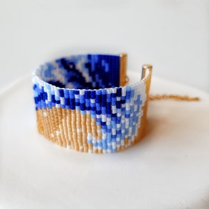 K&ouml;nnte beinhalten: Ein Armband aus Perlen mit einem blauen, wei&szlig;en und beigefarbenen Muster. Das Armband ist aus kleinen, bunten Perlen gefertigt und hat einen goldenen Verschluss.
