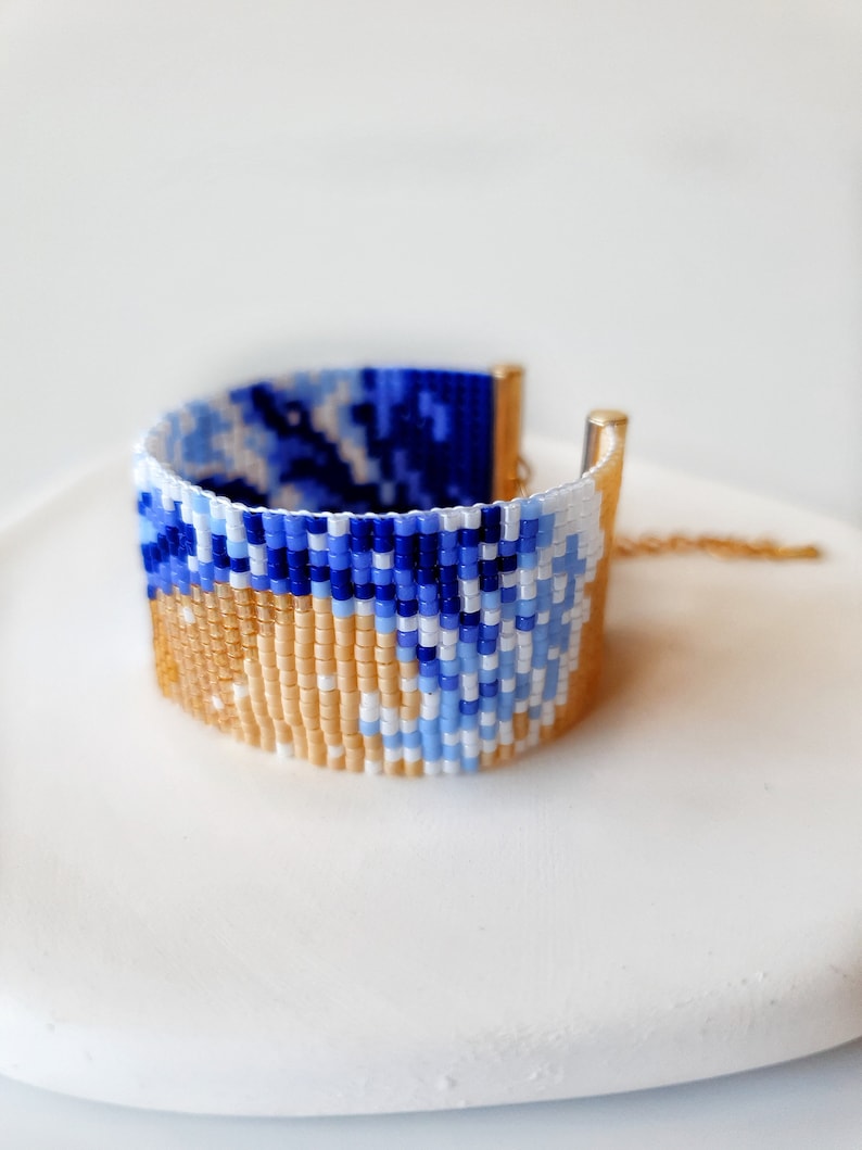 K&ouml;nnte beinhalten: Ein Armband aus Perlen mit einem blauen, wei&szlig;en und beigefarbenen Muster. Das Armband ist aus kleinen, bunten Perlen gefertigt und hat einen goldenen Verschluss.