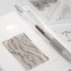 May include: A close-up of a silver and white beaded bracelet with a wavy pattern. The bracelet is lying on a white surface with a pen and a logo that says "AU Jewelry".