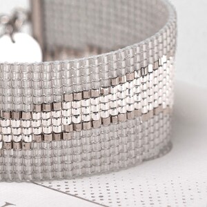 K&ouml;nnte beinhalten: Ein silbernes und wei&szlig;es Armband mit geometrischem Muster. Das Armband besteht aus kleinen, quadratischen Perlen, die zu einem einzigartigen Design verwoben sind.
