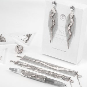 Könnte beinhalten: Ein Set aus silbernen Perlen-Schmuck, bestehend aus einem Paar Ohrringen, einem Armband, einem Ring und einem Stift. Die Ohrringe sind lang und baumelnd, das Armband ist breit und flach, und der Ring ist schlicht und elegant. Der Stift ist mit silbernen Perlen verziert und hat eine silberne Kappe. Der Schmuck ist in einer weißen Schachtel mit den Worten "Charming things inside" verpackt.