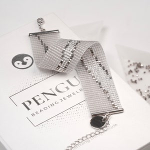 K&ouml;nnte beinhalten: Ein silbernes Perlenarmband mit einem silbernen Verschluss und wei&szlig;em Hintergrund. Das Armband hat einen silbernen Streifen, der in der Mitte verl&auml;uft. Das Armband befindet sich in einer Schachtel mit dem Aufdruck "PENGU Beading Jewelry".
