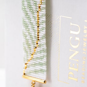 Pode incluir: Um bracelete de contas brancas e verdes com uma corrente e um fecho dourados. O bracelete está sobre um fundo branco com o texto "PENGU BEADING JEWELRY" em letras douradas.