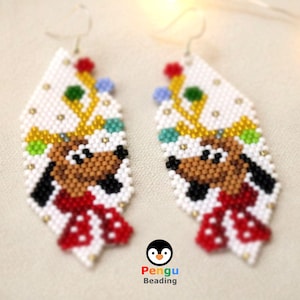Może przedstawiać: Para ręcznie robionych kolczyków z koralików z motywem renifera z kreskówki. Kolczyki mają białe tło z twarzą renifera, porożem i czerwoną kokardą. Kolczyki mają kształt rombu i srebrne haczyki.