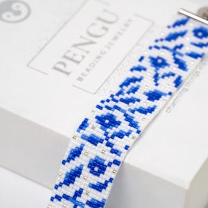 Pode incluir: Um bracelete de contas brancas e azuis com um fecho prateado. O bracelete está sobre uma caixa branca com o texto "PENGU Beading Jewelry" impresso nela.