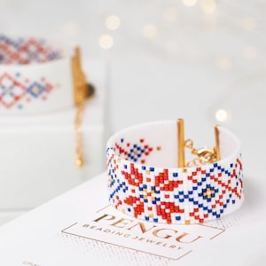 May include: White beaded bracelet with a red, blue, and gold geometric pattern. The bracelet has a gold clasp and is on a white surface with the text "PENGU BEADING JEWELRY".