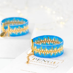 Peut inclure: Deux bracelets en perles bleues et dorées sur un fond blanc. Les bracelets présentent un motif géométrique et sont faits de petites perles. Les bracelets sont sur une surface blanche avec le texte "PENGU BEADING JEWELRY" imprimé sur la surface.