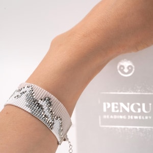 May include: A wide, beaded bracelet in white, grey, and silver tones. The bracelet features a geometric pattern and is worn on a wrist. The bracelet is made of small beads and has a silver clasp. The brand name "Pengu Beading Jewelry" is visible in the background.