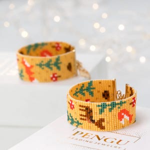Könnte beinhalten: Zwei gelbe Perlenarmbänder mit Waldmotiv. Die Armbänder zeigen einen Fuchs, ein Reh, Bäume und Pilze. Die Armbänder sind aus kleinen, bunten Perlen gefertigt und haben einen goldenen Kettenverschluss.