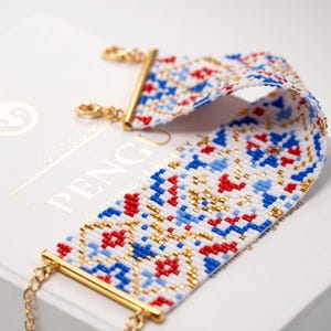 May include: A white beaded bracelet with a geometric pattern in red, blue, and gold. The bracelet has a gold clasp and is on a white box with the word "PENG" printed on it.