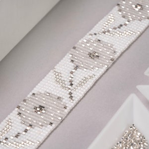 May include: A white and silver beaded bracelet with a repeating pattern of small squares. The bracelet is made with seed beads and is lying on a gray surface.