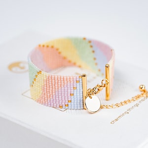 Pode incluir: Um bracelete de contas de arco-íris com um fecho dourado e um pequeno amuleto dourado. O bracelete está sobre uma superfície branca com o texto "charming things rock" impresso nele.