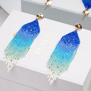 Könnte beinhalten: Ein Paar lange, blaue, grüne und weiße Perlen-Fransenohrringe mit goldenen Akzenten. Die Ohrringe sind im Ombre-Stil gehalten und haben ein abgestuftes Farbmuster.