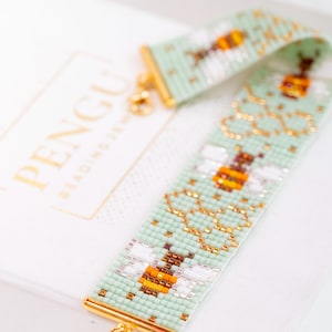 Puede incluir: Una pulsera ancha de cuentas con un fondo verde menta claro, decorada con patrones repetitivos de abejas amarillas y marrones y contornos de panal dorados. La pulsera está rematada con terminales de metal dorado y un cierre de mosquetón.