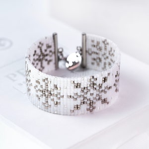 May include: White beaded bracelet with a snowflake design. The bracelet features a wide band of white beads with silver-colored accents. The bracelet has a silver-colored clasp and a small, round charm.
