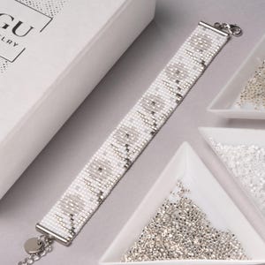 May include: A white and silver beaded bracelet with a silver clasp. The bracelet is on a white surface with a white box and small bowls of beads.