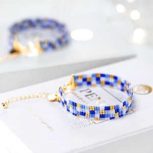 以下が含まれることがあります： 青と白のビーズブレスレットで、金色のアクセントが施されています。ブレスレットは市松模様になっており、小さな正方形のビーズで作られています。ブレスレットは、「Peace」という文字が印刷された白い表面の上に置かれています。