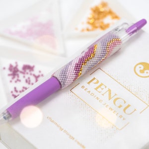Puede incluir: Un bolígrafo morado con un diseño de cuentas en tonos morados, blancos y dorados. El bolígrafo está sobre una caja blanca con el texto "PENGU Beading Jewelry" impreso en dorado.