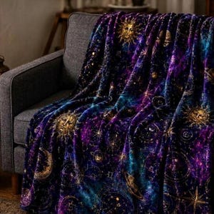 May include: A plush throw blanket featuring a deep purple and blue galaxy background with gold celestial motifs including suns, moons, planets, and astrological symbols scattered across the fabric, draped over a dark grey upholstered sofa.