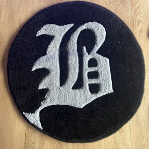 Puede incluir: Una alfombra redonda negra y blanca con una gran letra blanca "B" en una fuente estilizada.